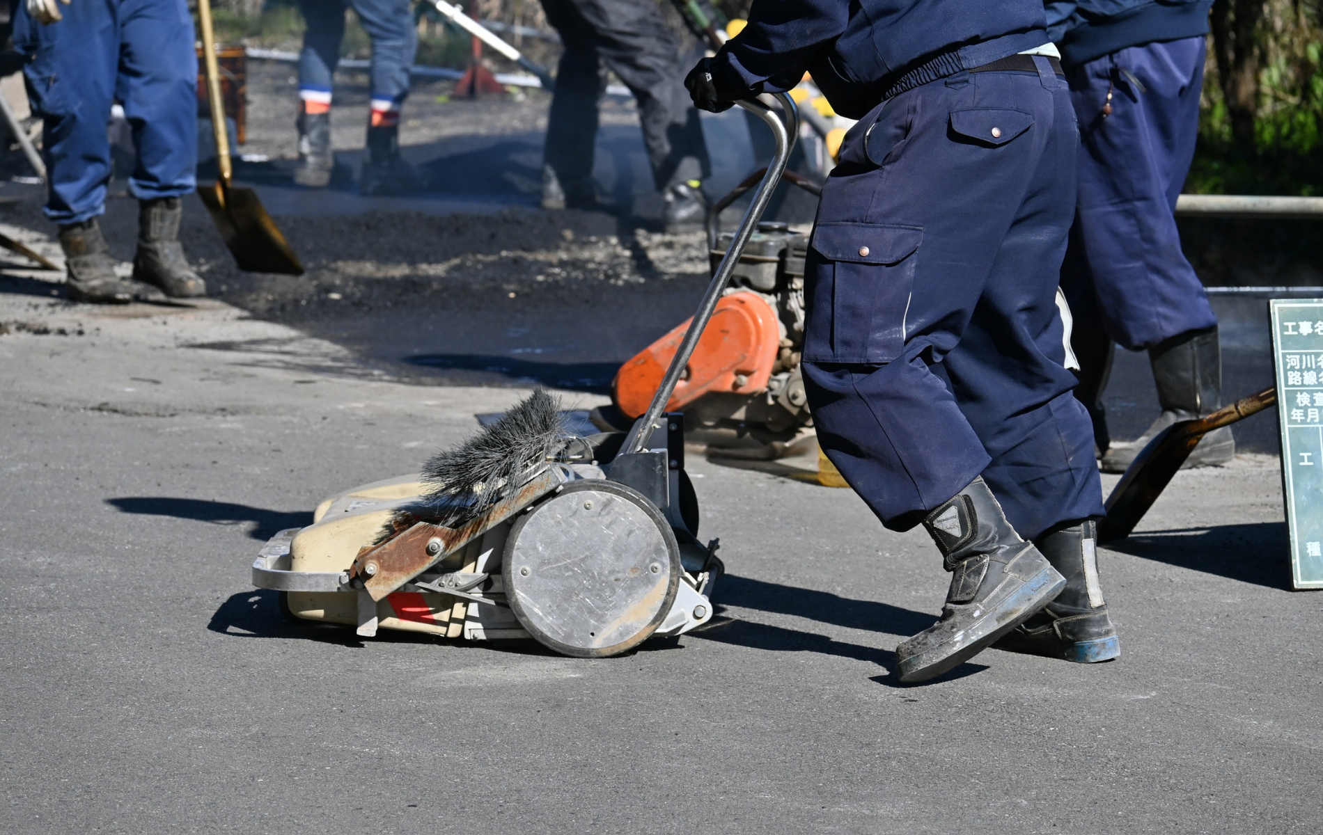 Workers paving road with asphalt, using a machine. Other workers sweep and assist.