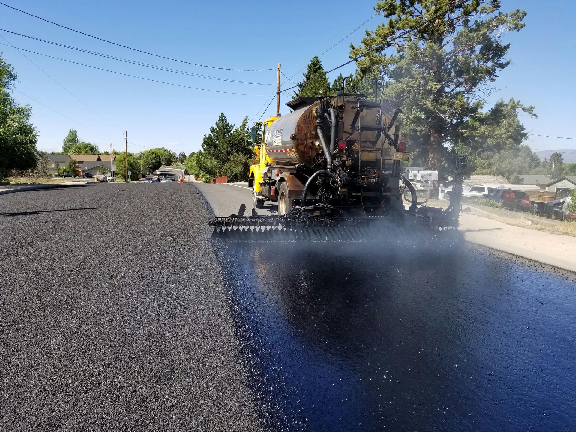 Road paving machine spraying asphalt on a street; sunny day, residential setting.