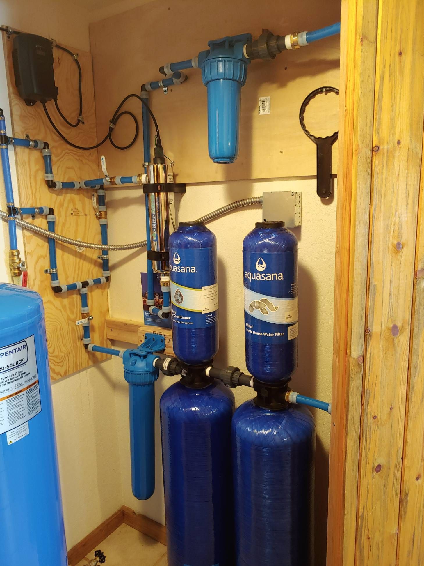 Water filtration system with blue tanks, pipes, and filters, mounted on a wooden wall.