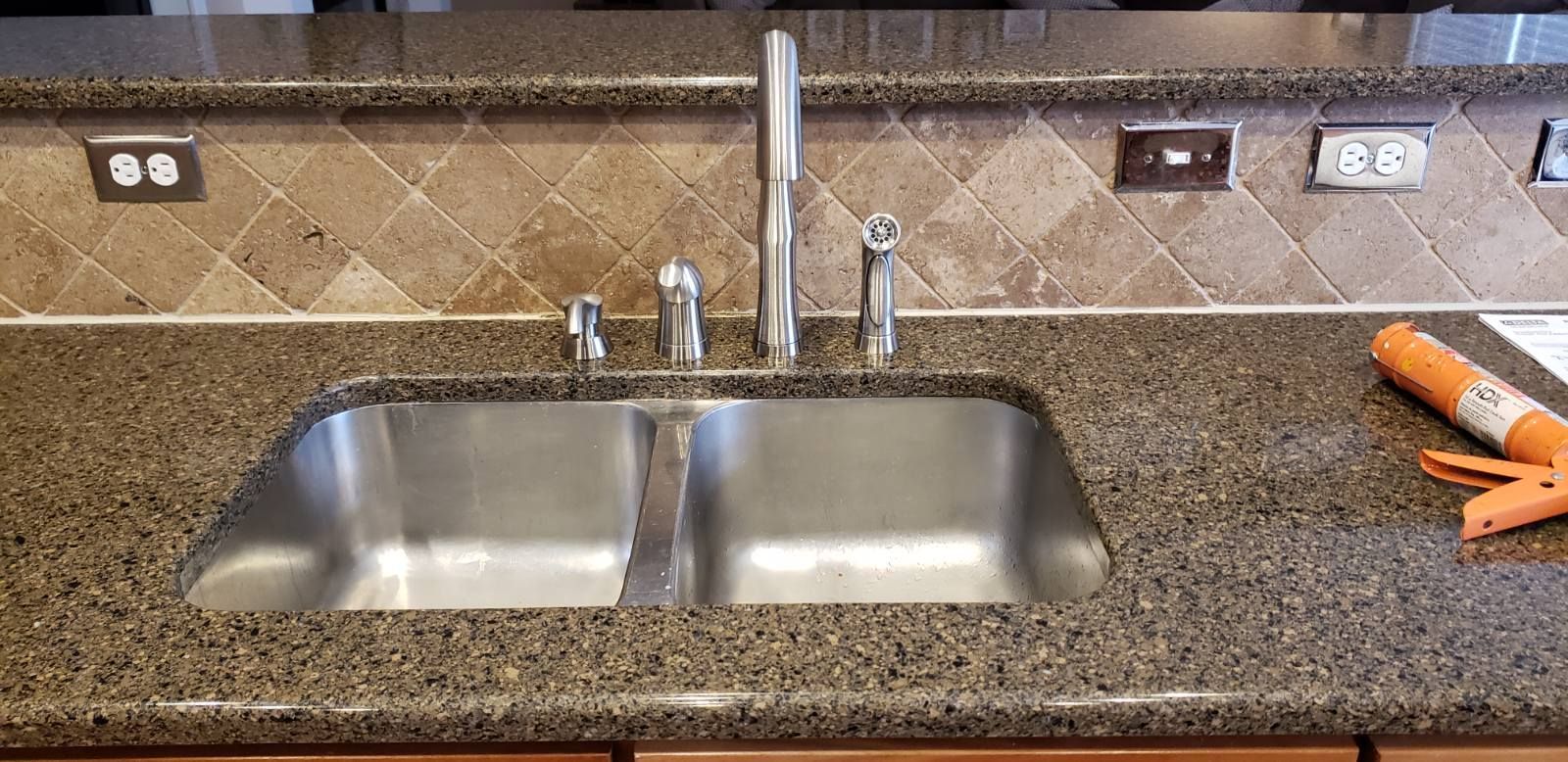 A stainless steel sink is centered in a mottled brown countertop, next to an orange caulk gun.