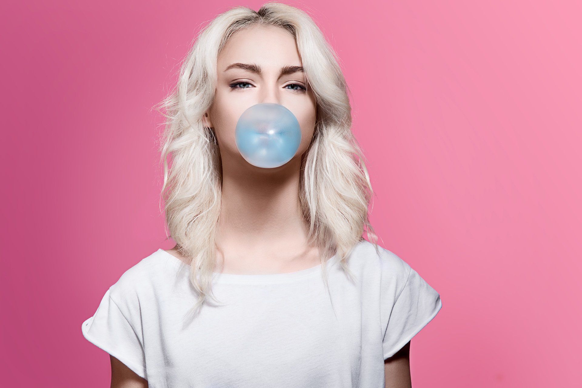 A woman is blowing a bubble of gum in front of a pink background.