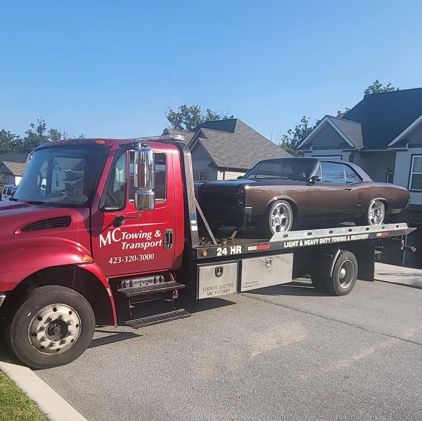 A red MC Towing & Transport flatbed truck carries a brown vintage muscle car on a residential street.