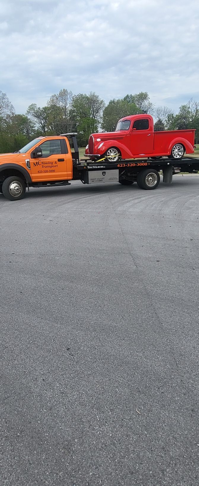 An orange flatbed tow truck transporting a vintage red pickup truck across a paved lot.