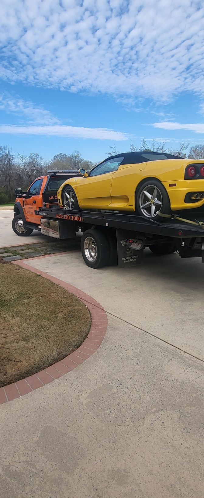 A yellow Ferrari convertible loaded onto the back of an orange flatbed tow truck on a driveway under a cloudy sky.