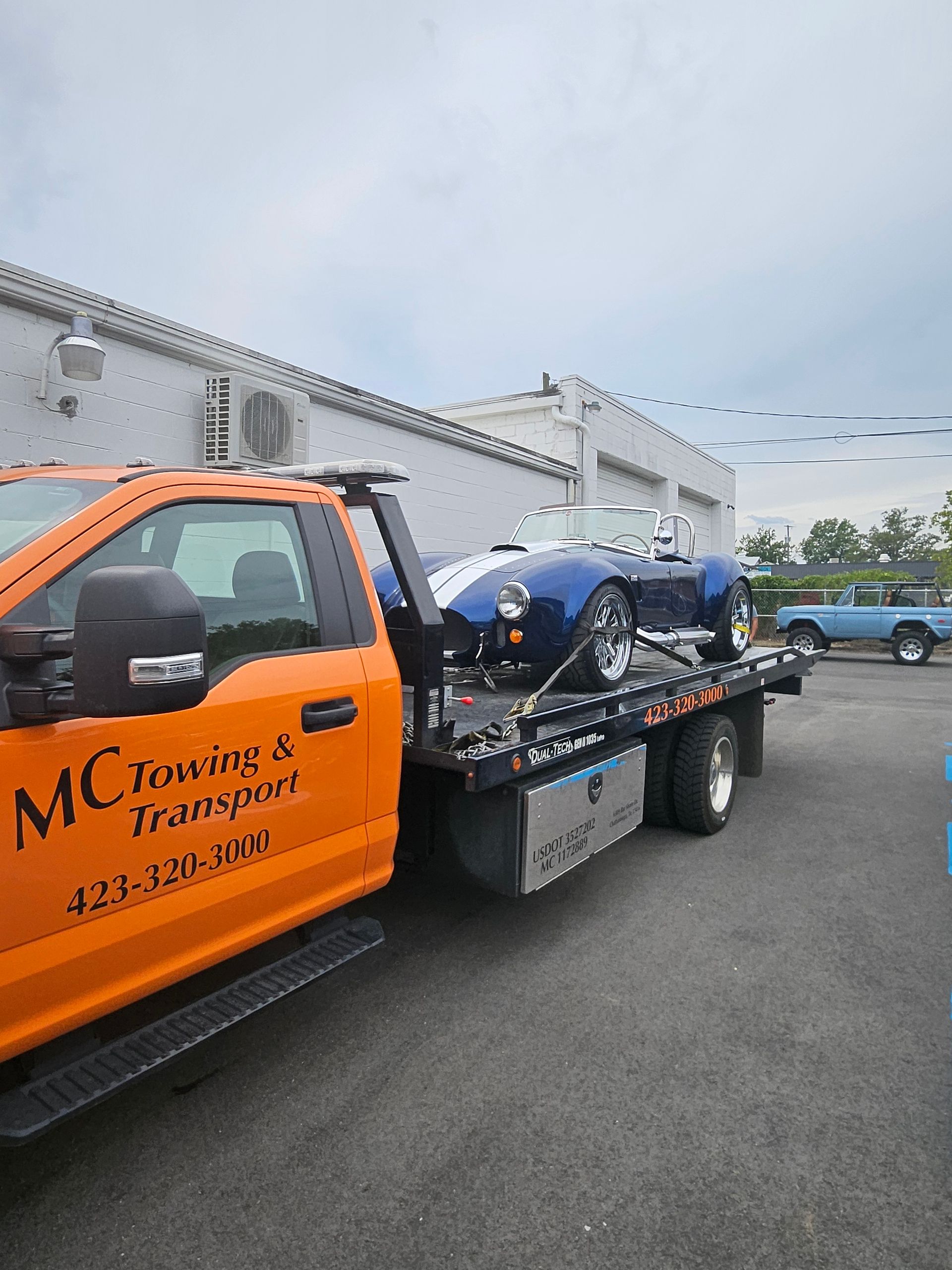 An orange MC Towing & Transport flatbed truck carries a dark blue convertible sports car with white racing stripes.