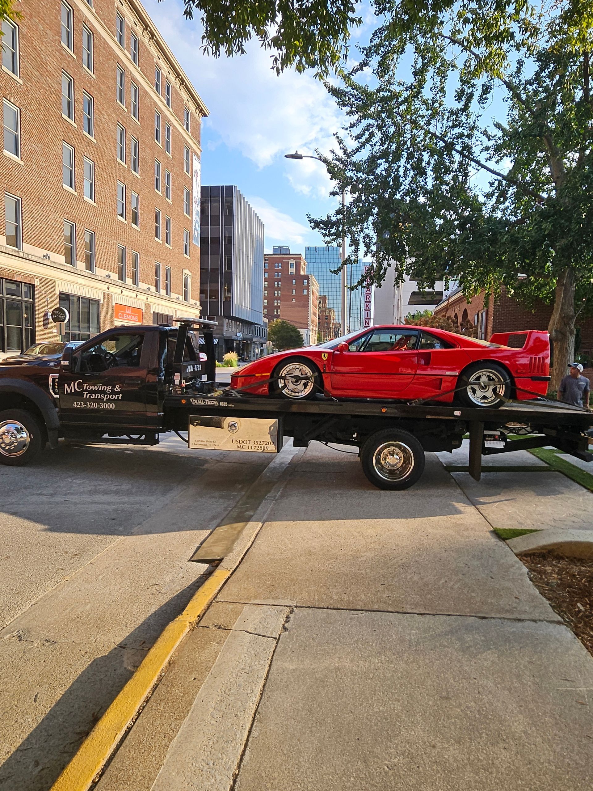 A bright red Ferrari F40 sports car being transported on the flatbed of a tow truck on a sunny city street.
