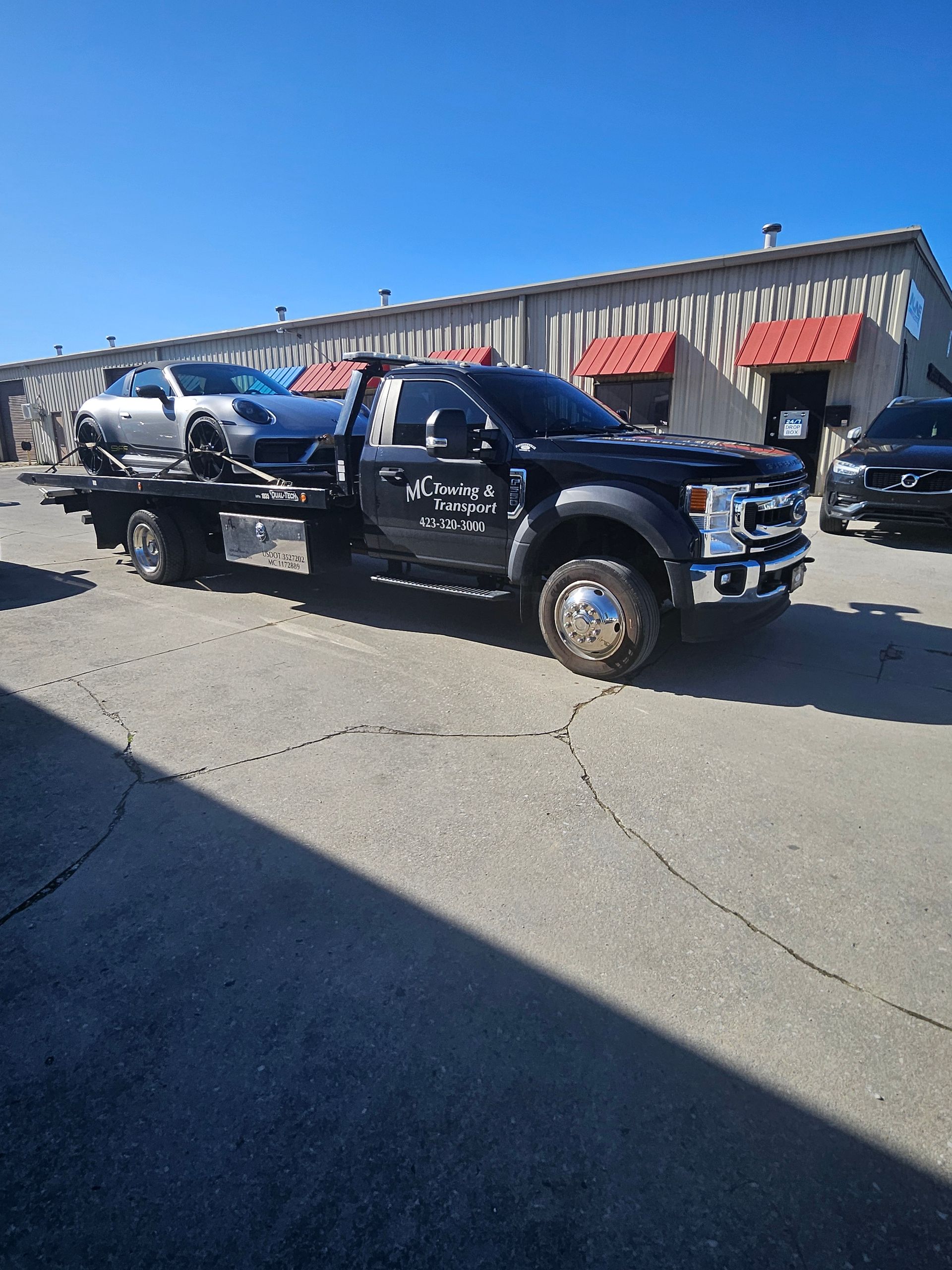 A black tow truck carrying a silver Porsche parked in front of a tan building under a clear blue sky.