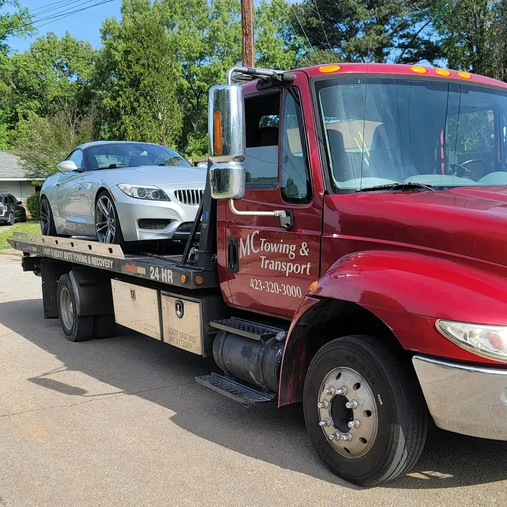 A damaged silver car being towed by a red tow truck on a residential street. Bright, sunny day.