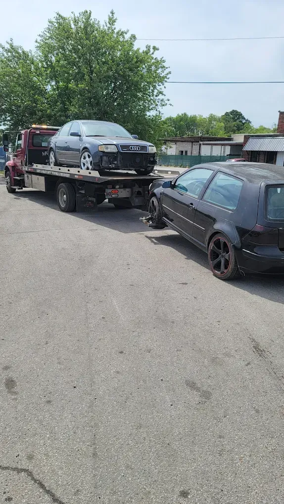 A flatbed tow truck carries a grey car, while a black hatchback is hooked and being towed behind it on an asphalt lot.