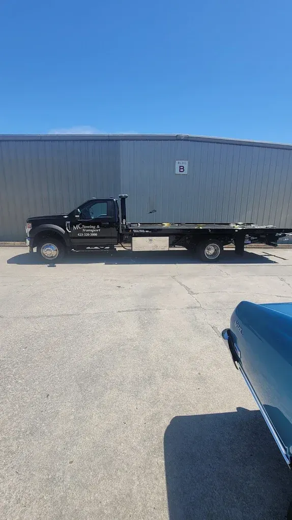 A black flatbed truck parked in front of a long metal industrial building on a sunny day.