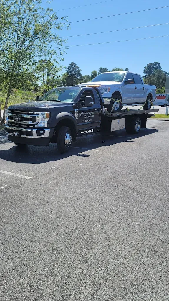 A black flatbed tow truck in a parking lot, transporting a white pickup truck.