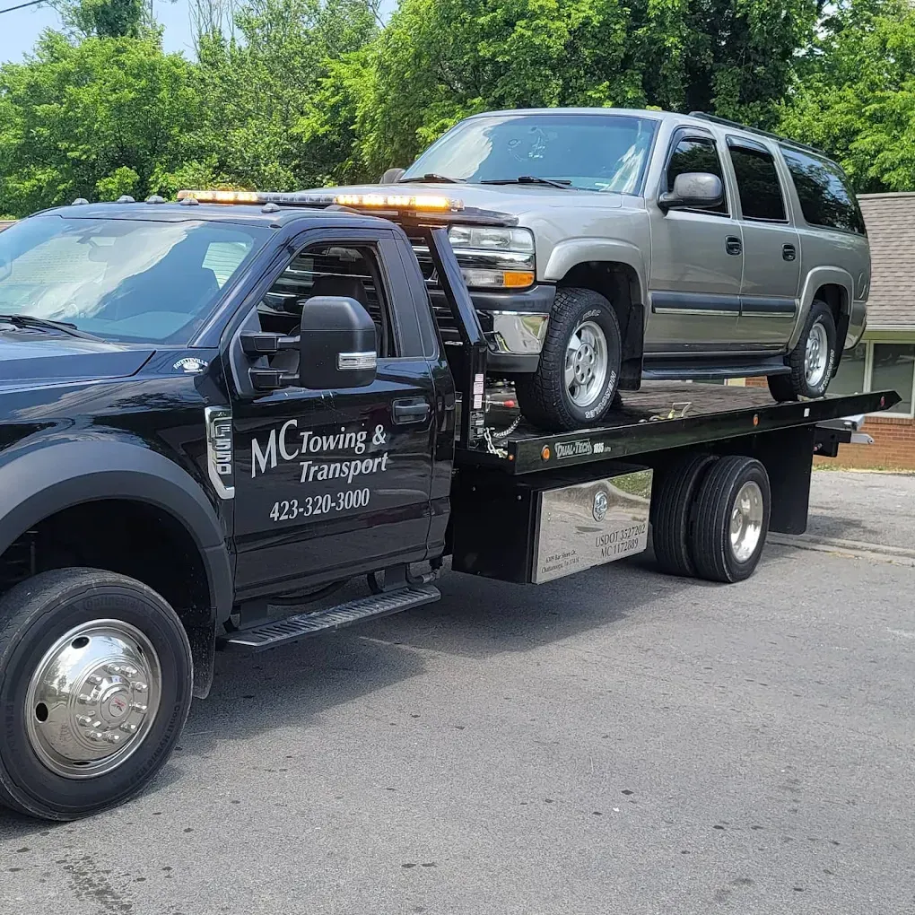 A black MC Towing & Transport flatbed truck hauling a silver SUV on a sunny day.