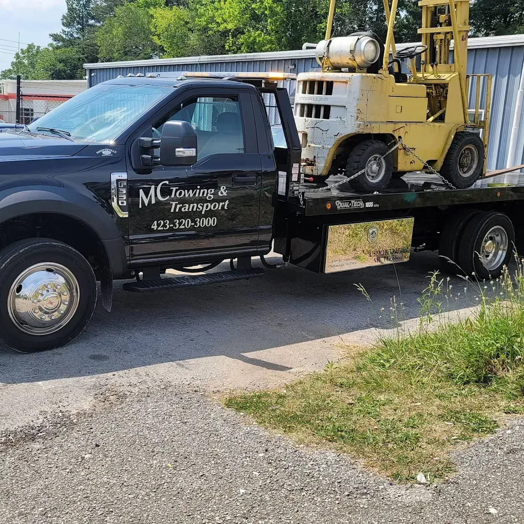 A black MC Towing & Transport flatbed truck is parked, carrying a yellow industrial forklift on its bed.