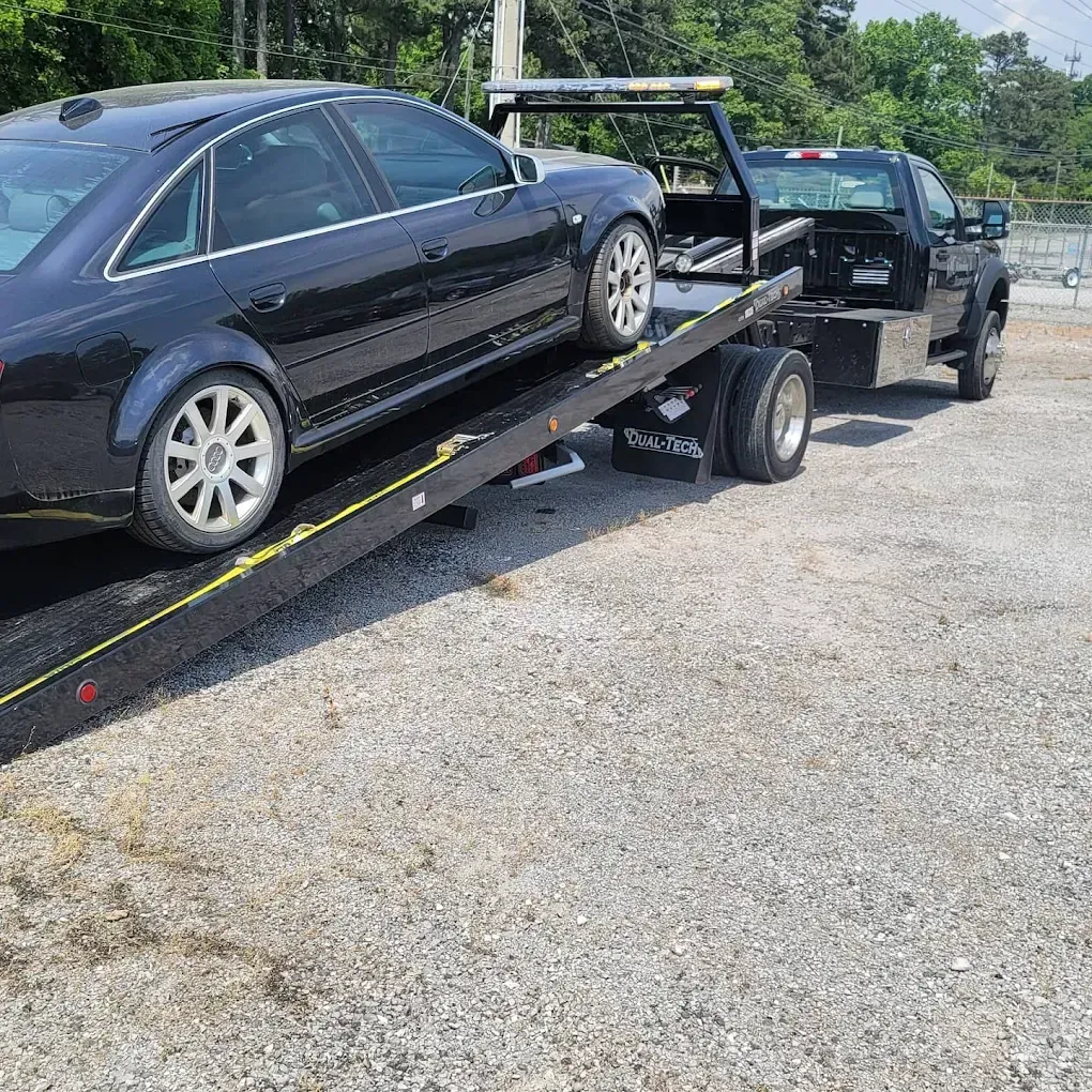 A black sedan is being loaded onto the flatbed of a black tow truck parked on a gravel lot under a bright sky.