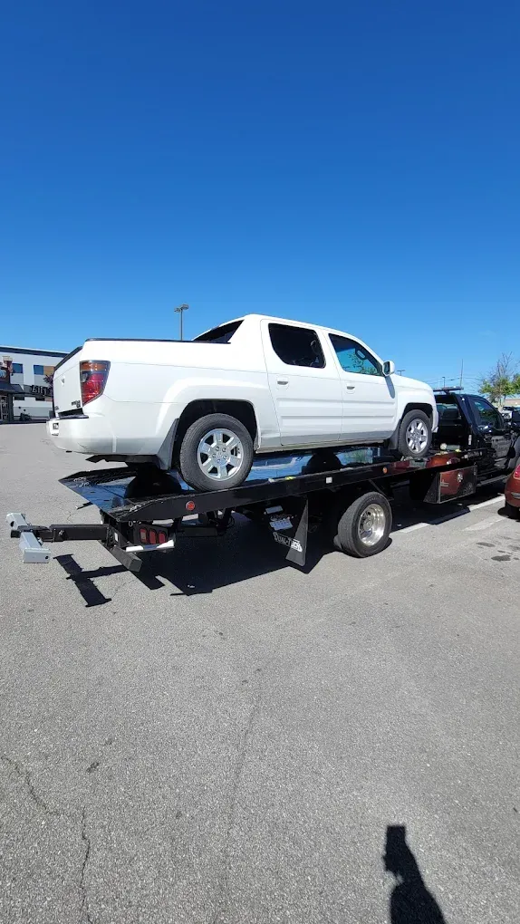 A white Honda Ridgeline pickup truck being transported on a flatbed tow truck in an outdoor parking lot.