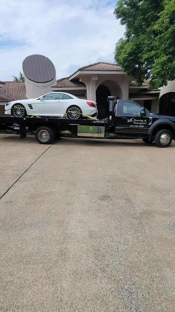 A white luxury convertible car secured on the flatbed of a black tow truck parked in front of a suburban home.