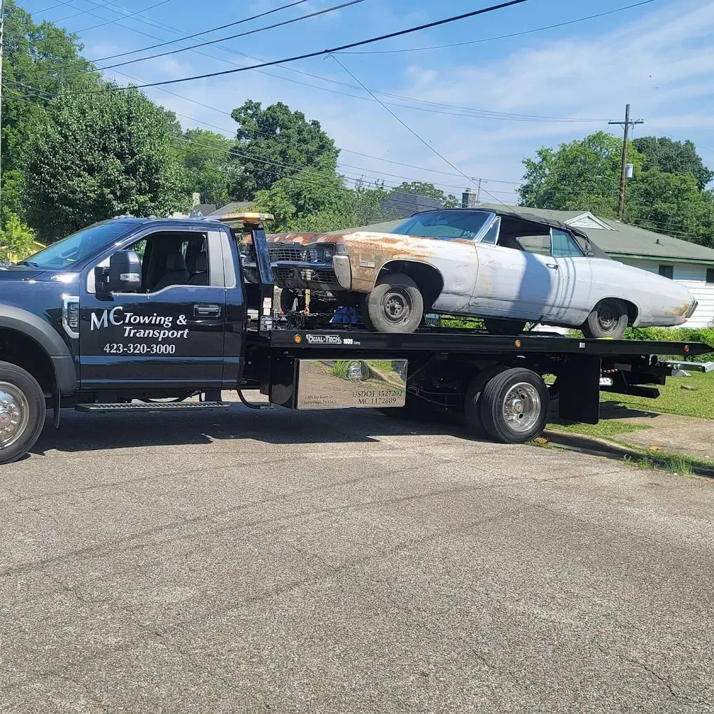 A dark-colored flatbed tow truck carries a vintage, light-colored convertible car on a sunny day.