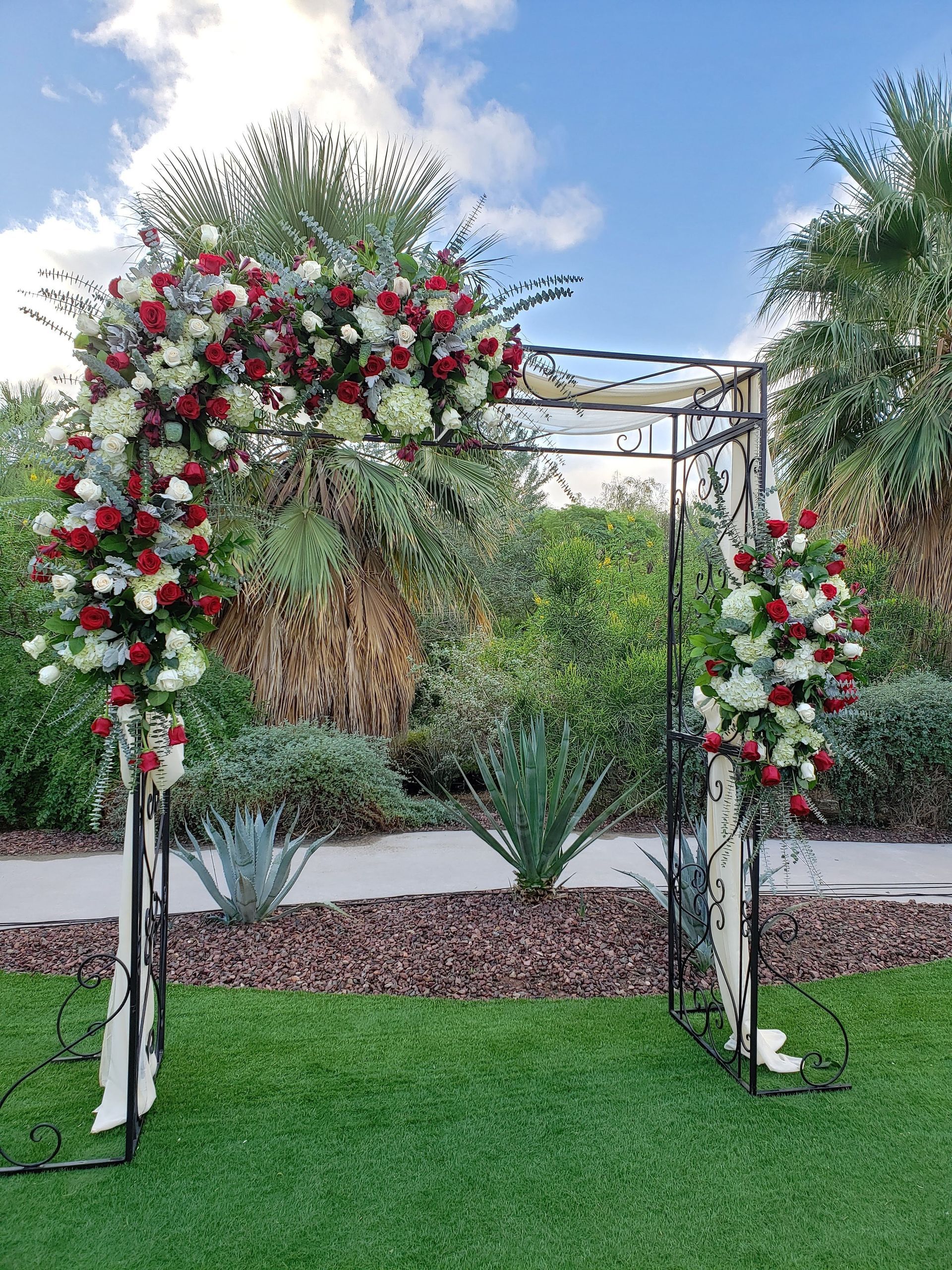 A wedding arch decorated with red and white flowers is sitting on top of a lush green field.