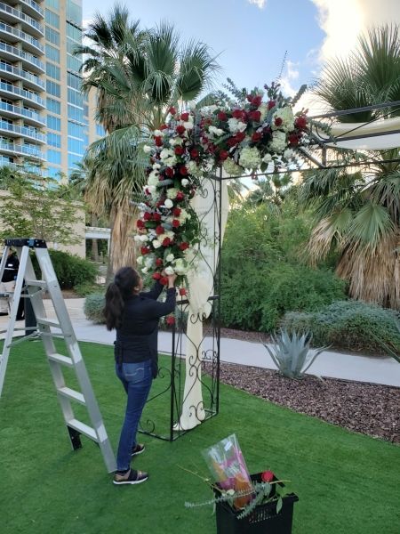 A woman is standing on a ladder decorating a wedding arch with flowers.