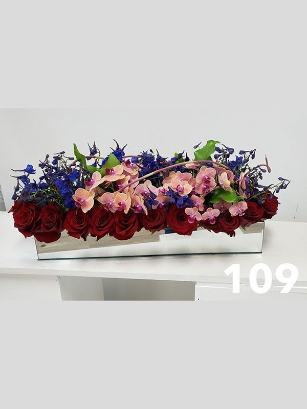 A long vase filled with red roses and blue flowers on a table.