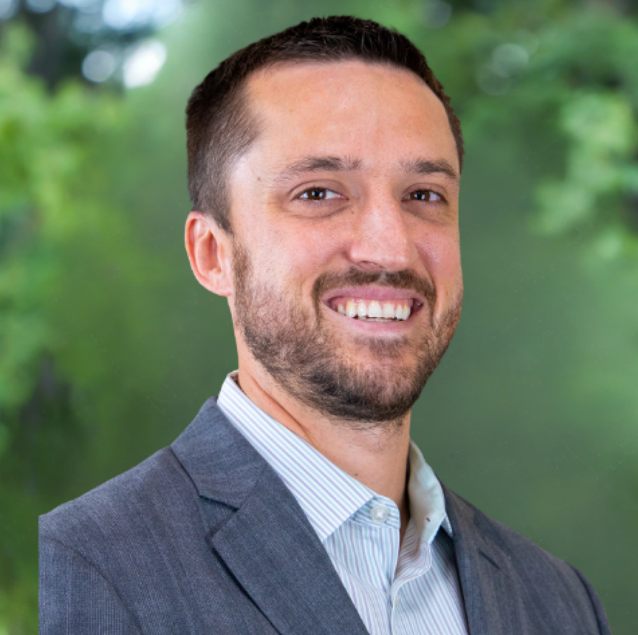 A man in a grey suit and light blue striped button-down shirt smiling against a blurred green outdoor background.