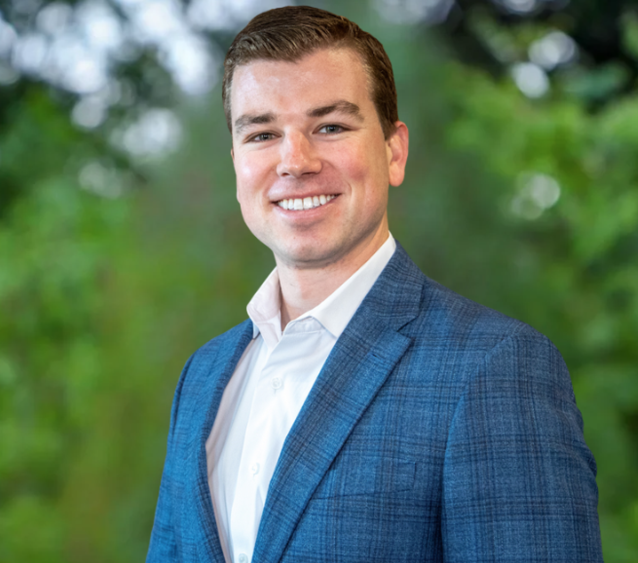 A person wearing a blue textured blazer and white collared shirt smiles against a soft-focus green leafy background.