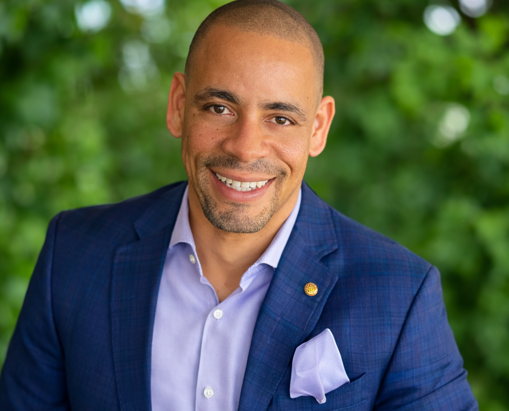 Smiling man wearing a blue blazer and light purple dress shirt with a pocket square, against a green leafy background.