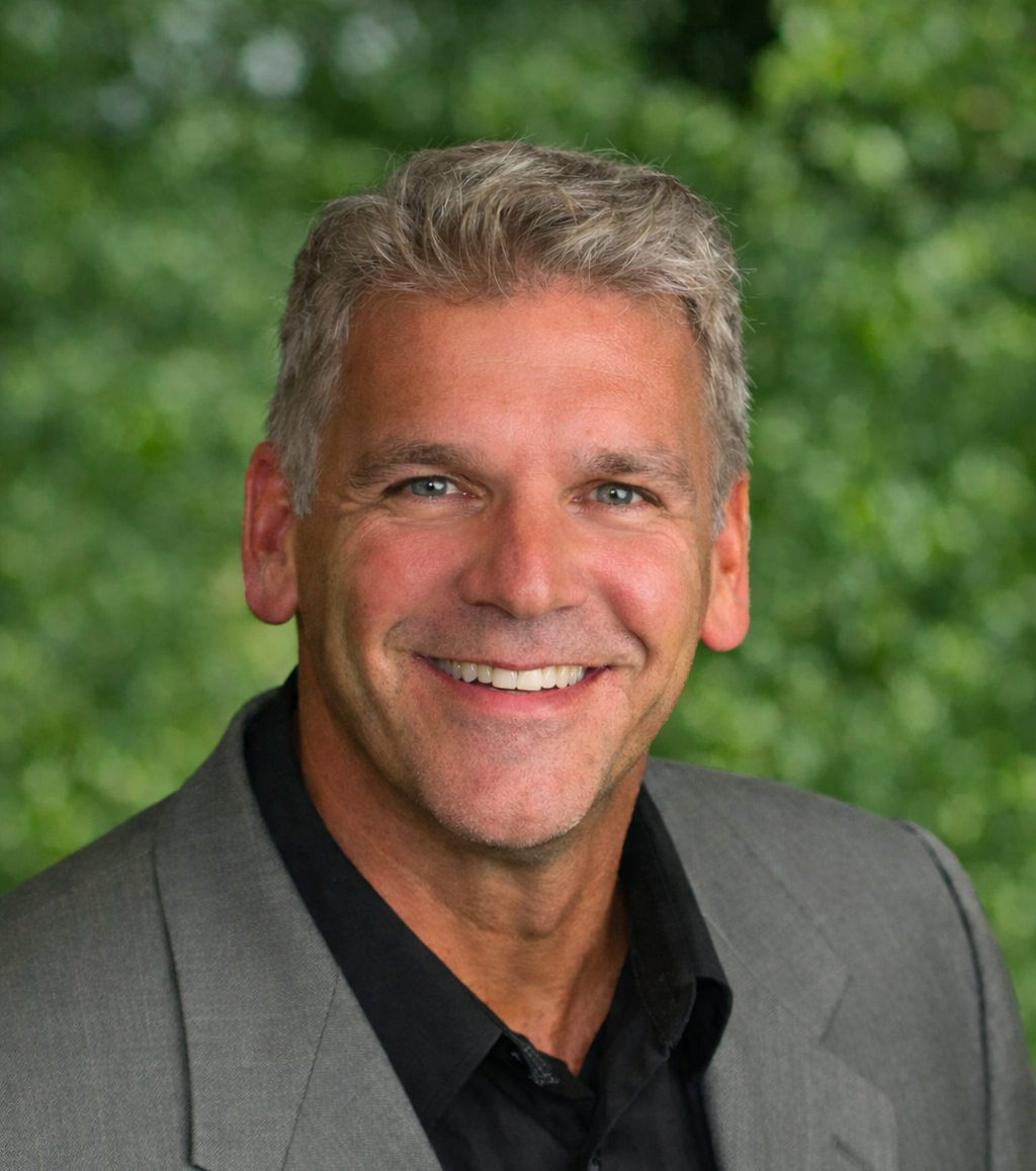 A smiling professional in a gray blazer and black shirt, positioned against a soft-focus green leafy background.