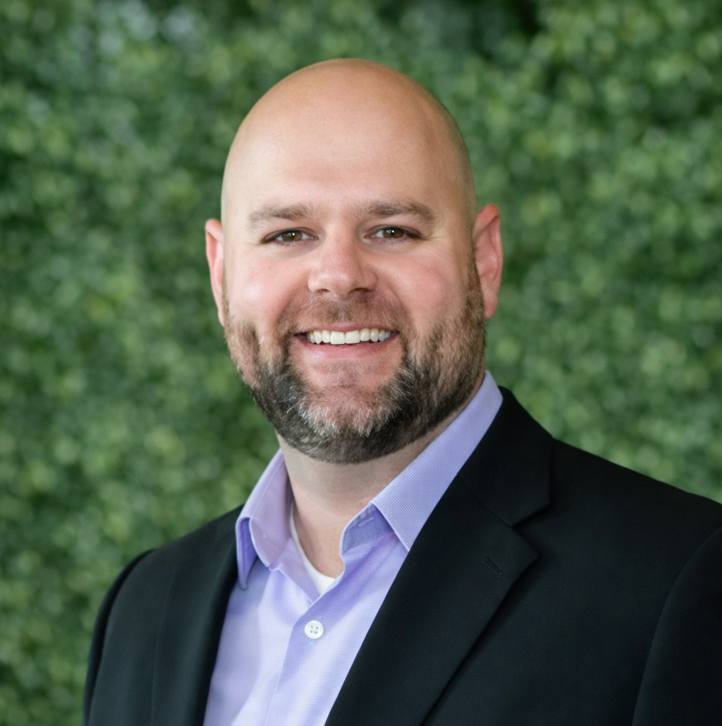 A smiling person with a beard, wearing a light purple shirt and a black blazer against a blurred green hedge background.