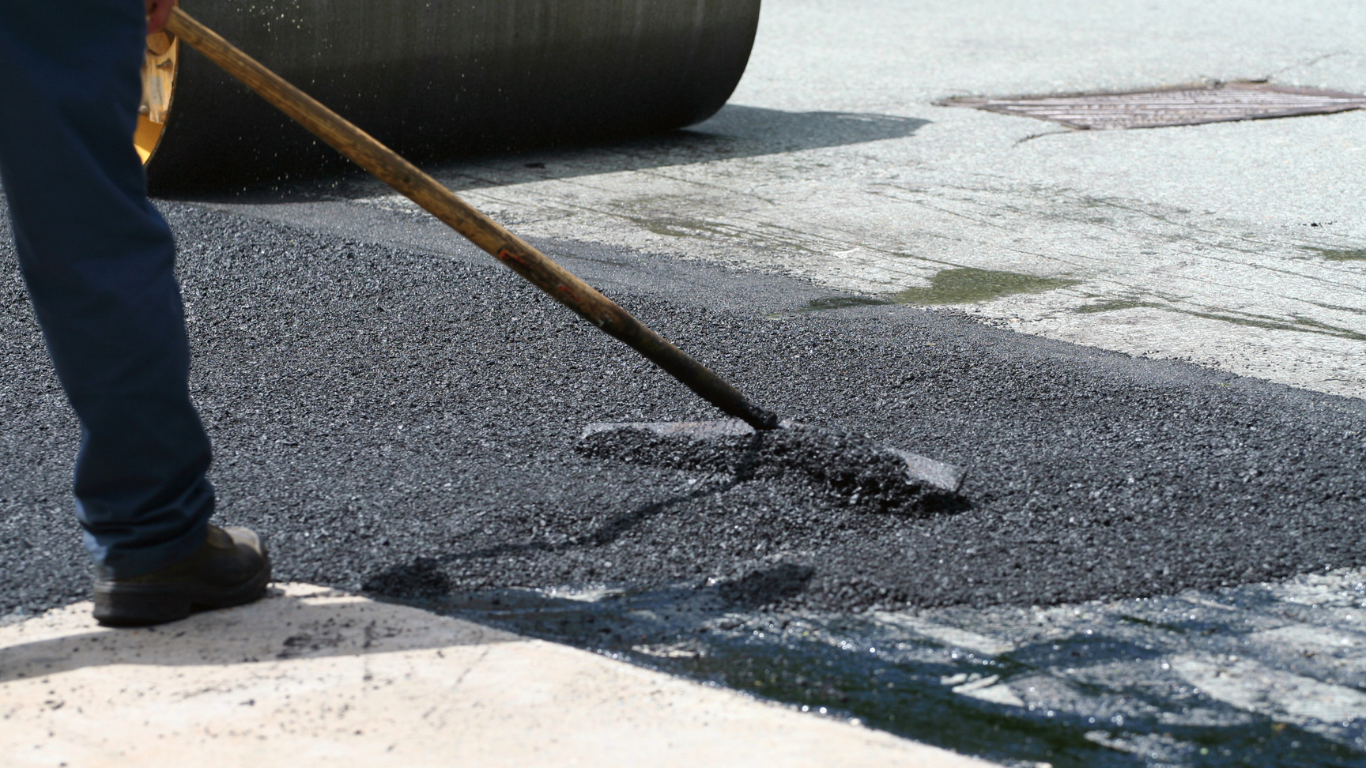 Person rakes hot asphalt, paving a road. Dark asphalt, tan edge.