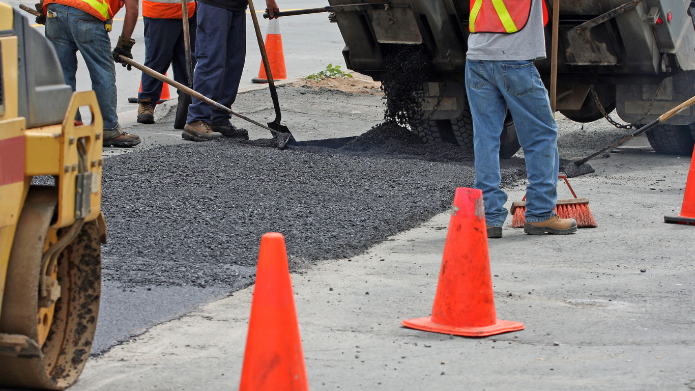 Road crew paving a street with asphalt; cones, roller, and workers in safety vests present.