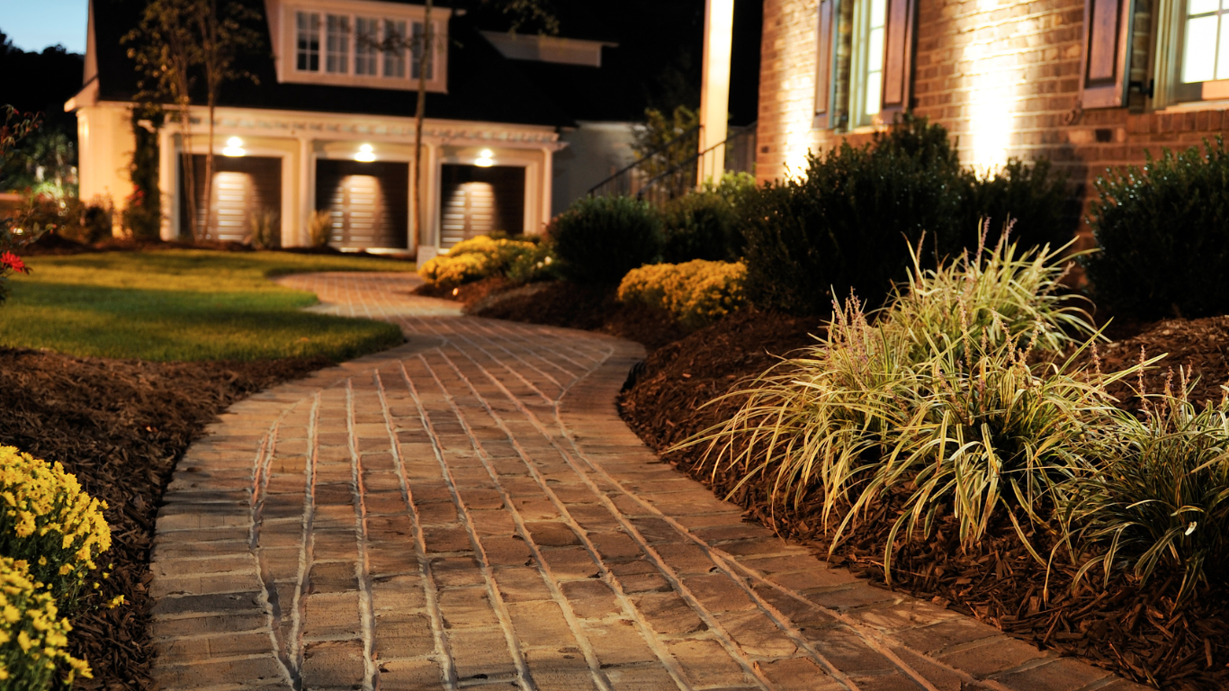 Brick walkway leads to a house at night, lit by landscaping lights, bordered by bushes and grass.