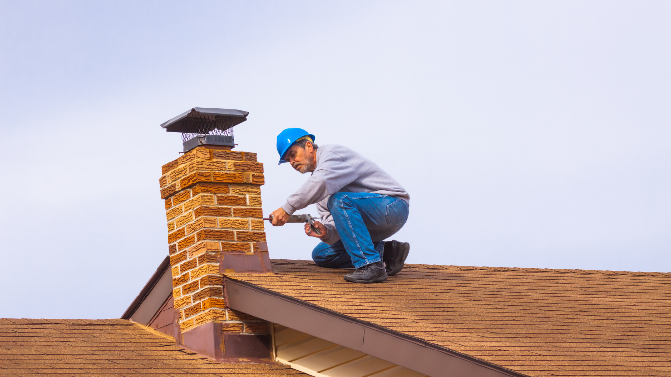 Man in hard hat on roof, repairing brick chimney with tools, brown roof, blue sky.