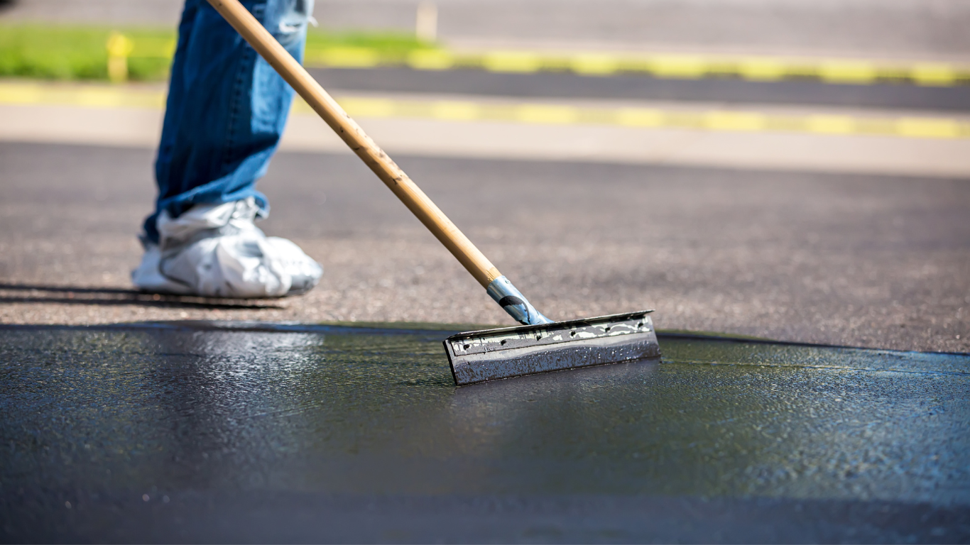 A person is using a broom to spread blacktop asphalt on the ground.