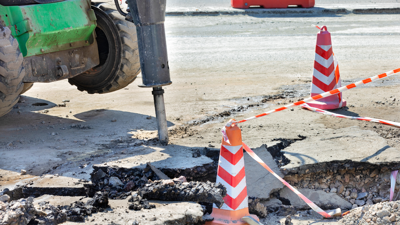 Jackhammer breaking up asphalt road, with cones and caution tape.