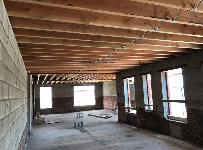 Interior view of a room under construction: wood ceiling beams, cinder block walls, windows, and a concrete floor.