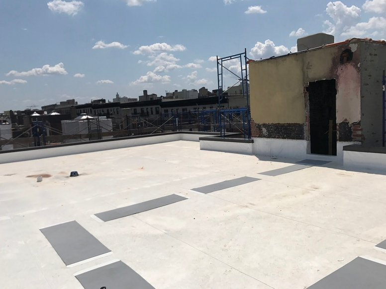 Flat, white rooftop with gray rectangles; a weathered building, scaffolding, and cityscape in the distance under a blue sky.