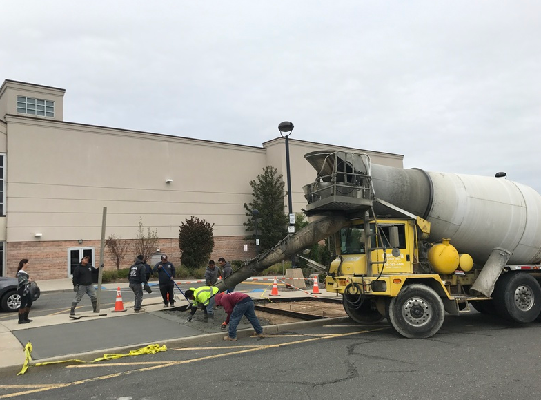 A yellow cement truck pours concrete onto a road; workers in reflective vests guide the pour.
