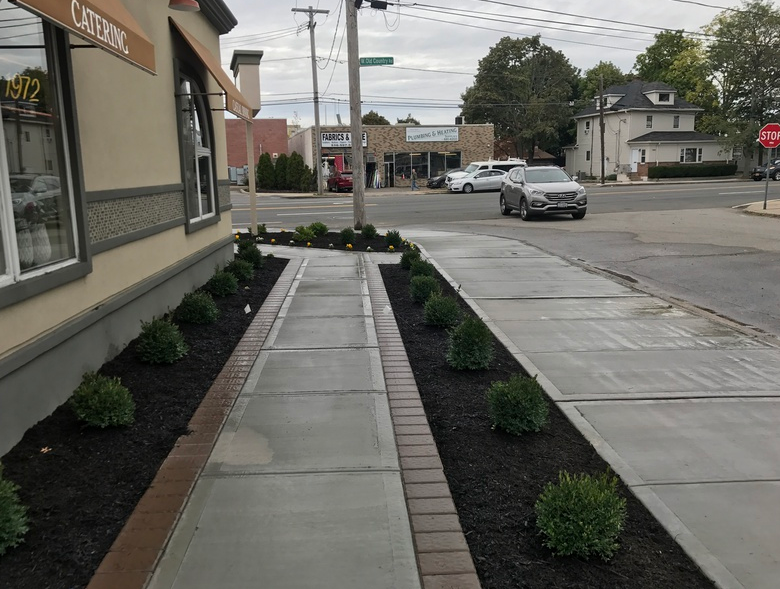 Sidewalk with planters of shrubs, next to a building with a sign, and street with cars.