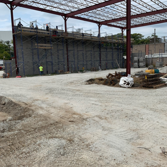Construction site with a large gray wall under a red steel frame, workers visible.
