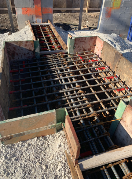 Concrete foundation with rebar grid, wooden forms, and orange supports. Construction site setting.
