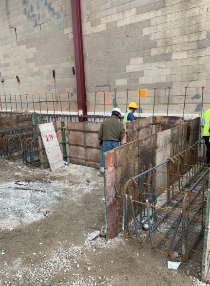 Construction workers building a concrete form with rebar, set against a brick wall.