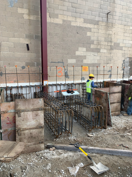Construction site with workers, rebar, concrete forms against a brick wall.