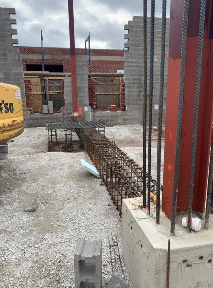 Construction site with steel beams, concrete, and rebar, in front of cinder block walls.