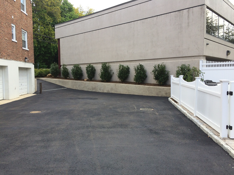 Asphalt driveway next to building with row of green bushes, white fence and garage door.