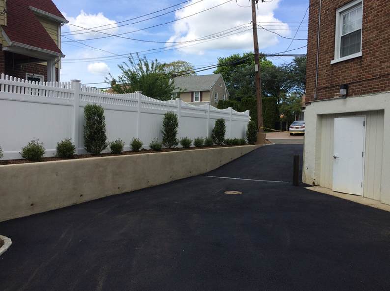 Asphalt driveway next to a white fence and concrete wall, with small green bushes and a garage.