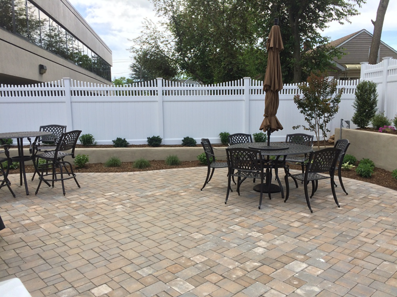 Outdoor patio with tables and chairs, surrounded by a white picket fence, trees, and a brick-paved ground.