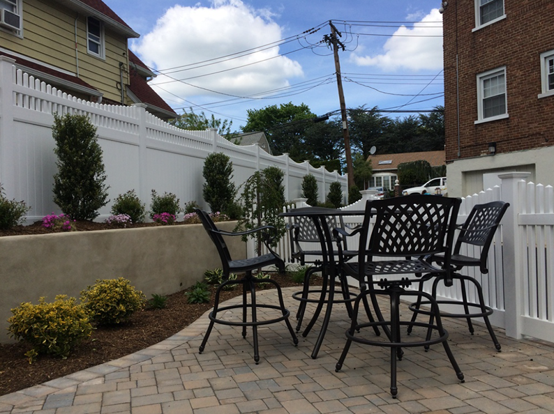 Patio with black table and chairs, white fence, and landscaping under a partly cloudy sky.