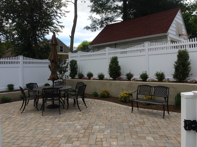 Patio with dark table/chairs and bench, white fence, red-roofed house, trees and landscaping.