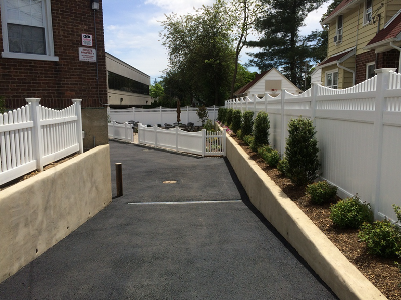 Driveway with white picket fence, brown mulch, and green bushes against a concrete wall and brick building.